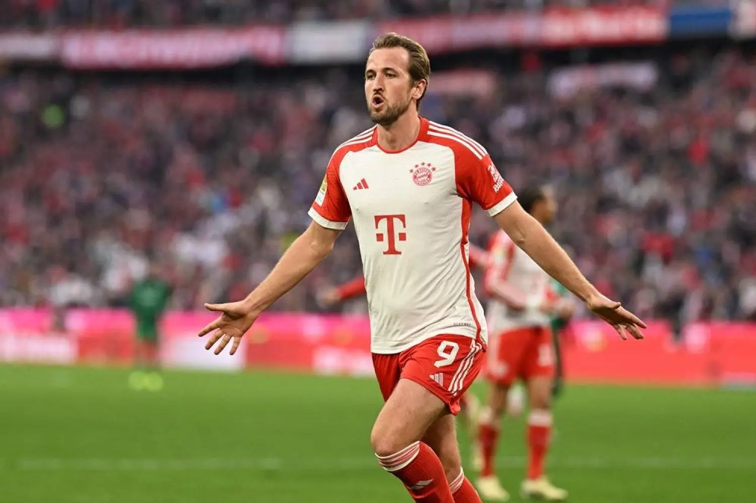 Harry Kane celebrating in a white and red Bayern Munich kit with arms outstretched on a football pitch