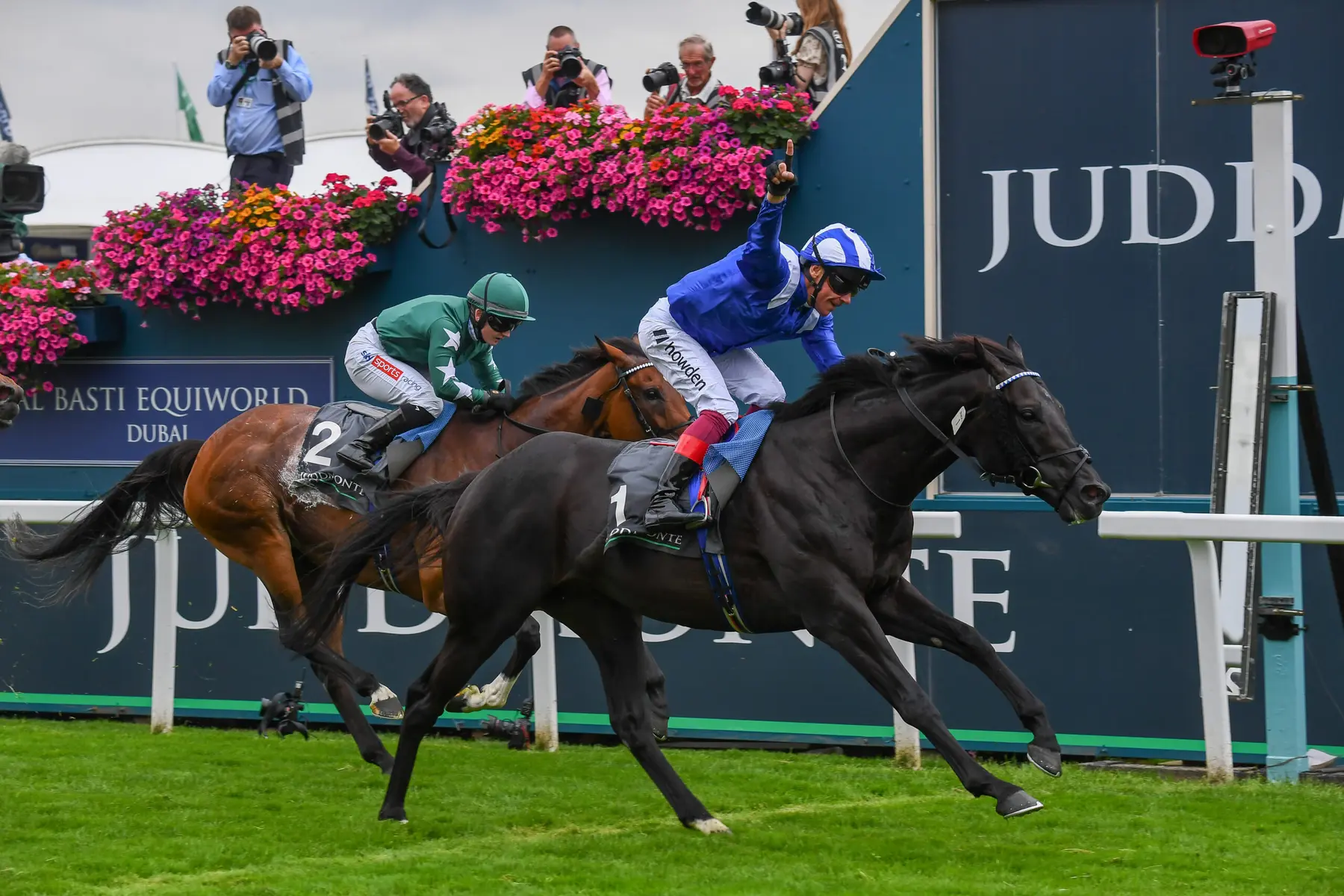 Two racehorses and jockeys galloping past a Juddmonte branded wall at a horse racing event