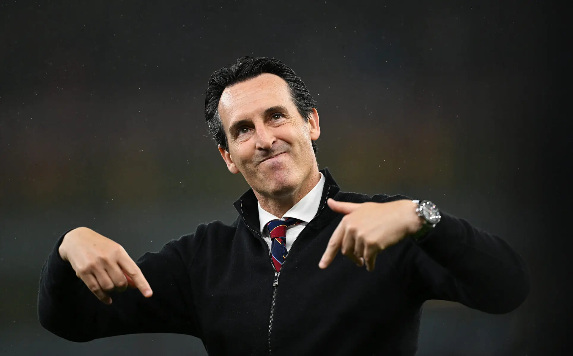 Unai Emery wearing a black zip-up jacket and striped tie, pointing downwards with both hands
