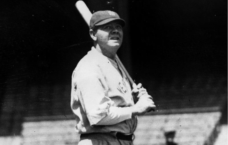 Babe Ruth holds a bat and looks at the stands.