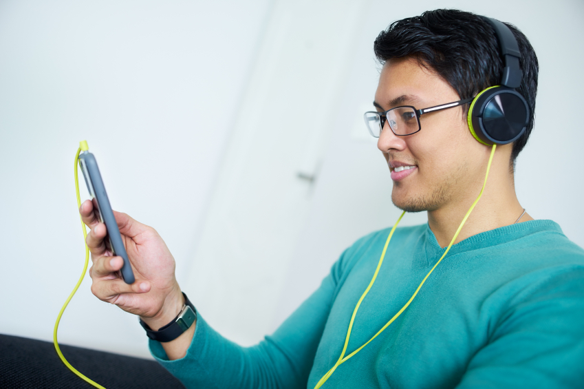 Chinese man relaxes on sofa and watches podcast on mobile phone, listening with green big earphones