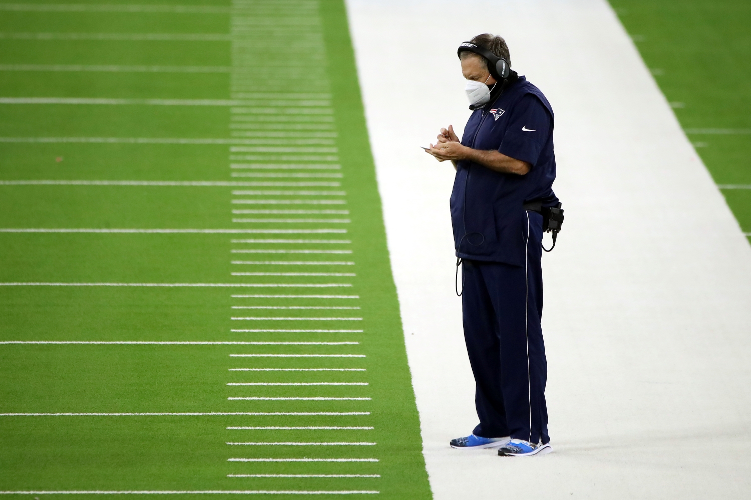 New England Patriots head coach Bill Belichick writes down notes while standing on the sideline.