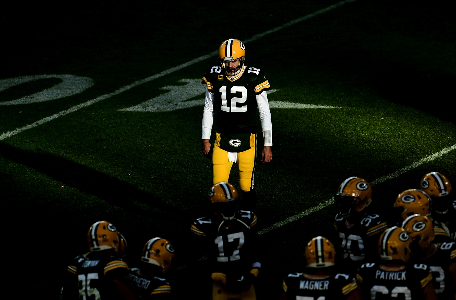 Green Bay Packers quarterback Aaron Rodgers stands a few feet away from his teammates as they huddle.