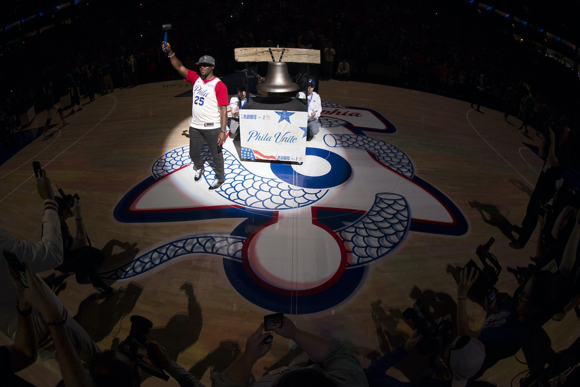 Jimmy Rollins rings the bell ahead of a Philadelphia 76ers playoff game, with the team's snake logo visible on the floor.