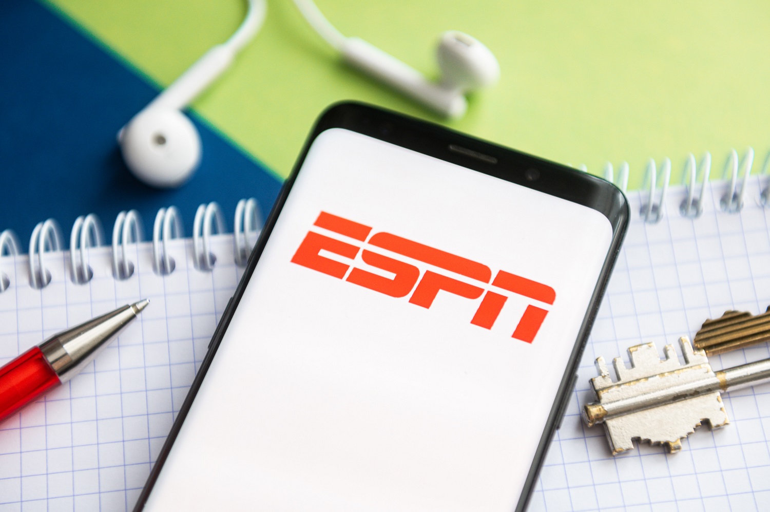 In this photo illustration, a ESPN logo seen displayed on a smartphone with a pen, key, book and headsets in the background.