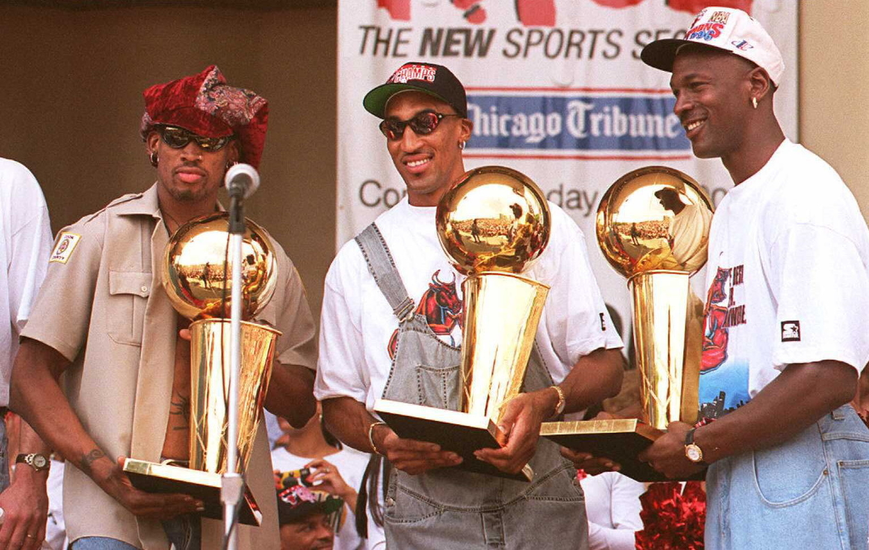 Scottie Pippen centering Dennis Rodman and Michael Jordan while holding up three Larry O'Brien trophies.