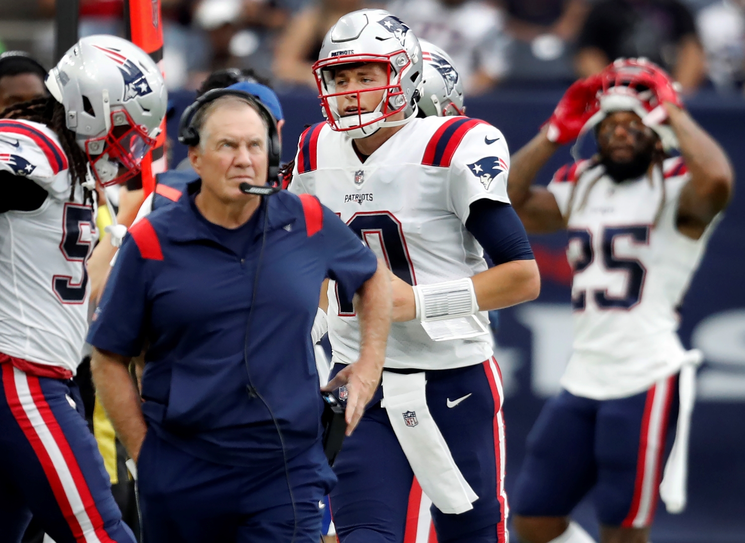 New England Patriots quarterback Mac Jones heads to the sidelines after throwing an interception while Bill Belichick looks up at the replay.