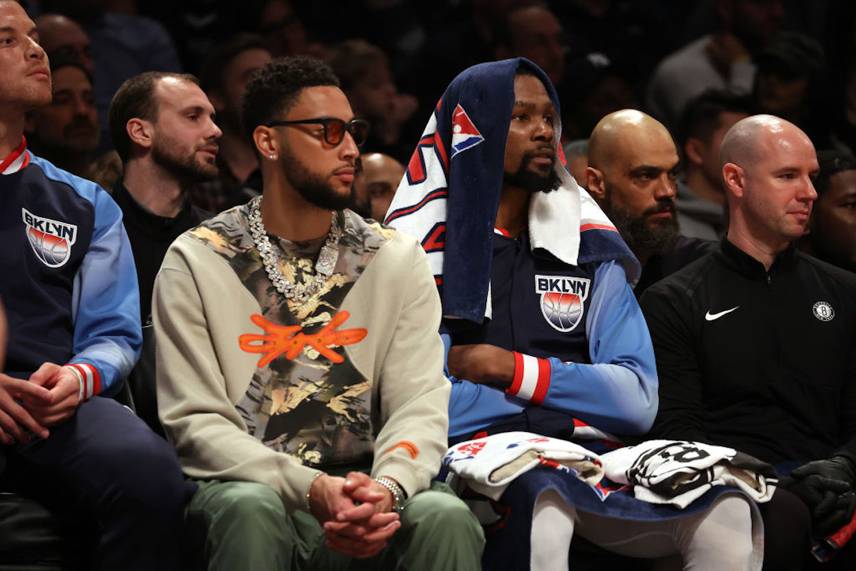 Injured Brooklyn Nets guard Ben Simmons (L) sits on the bench in street clothes.