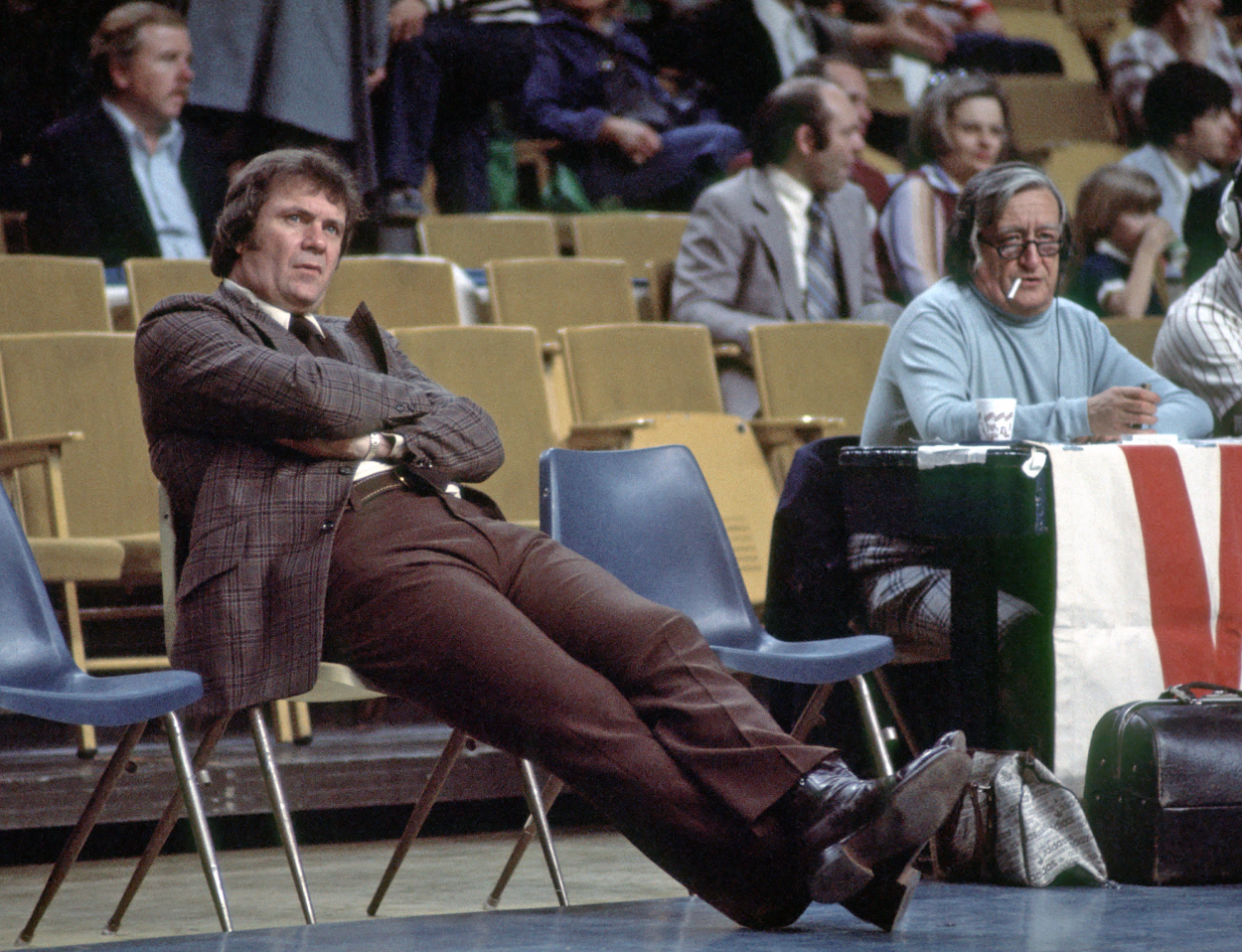 Head coach Tom Heinsohn of the Boston Celtics sits on a chair on the sideline before a game against the Buffalo Braves as Celtics radio announcer Johnny Most looks on.