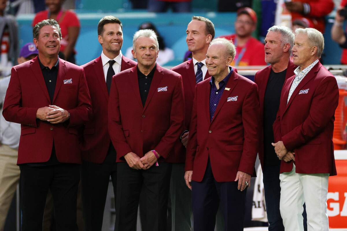 Dan Marino, Tom Brady, Joe Montana, Peyton Manning, Roger Staubach, Brett Favre, and John Elway stand on the field in red jackets at NFL 100 All-Time Team honors