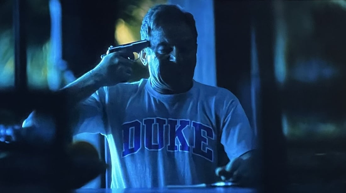 A man puts a gun to his head in a Duke shirt.