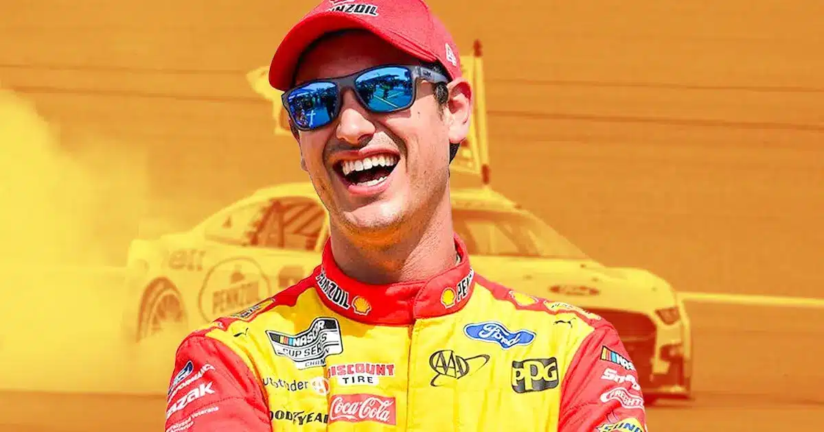 Smiling NASCAR driver Joey Logano in a yellow race suit with his car blurred in the background.