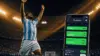 Soccer player in Argentina jersey celebrating goal with sports betting app in foreground at stadium