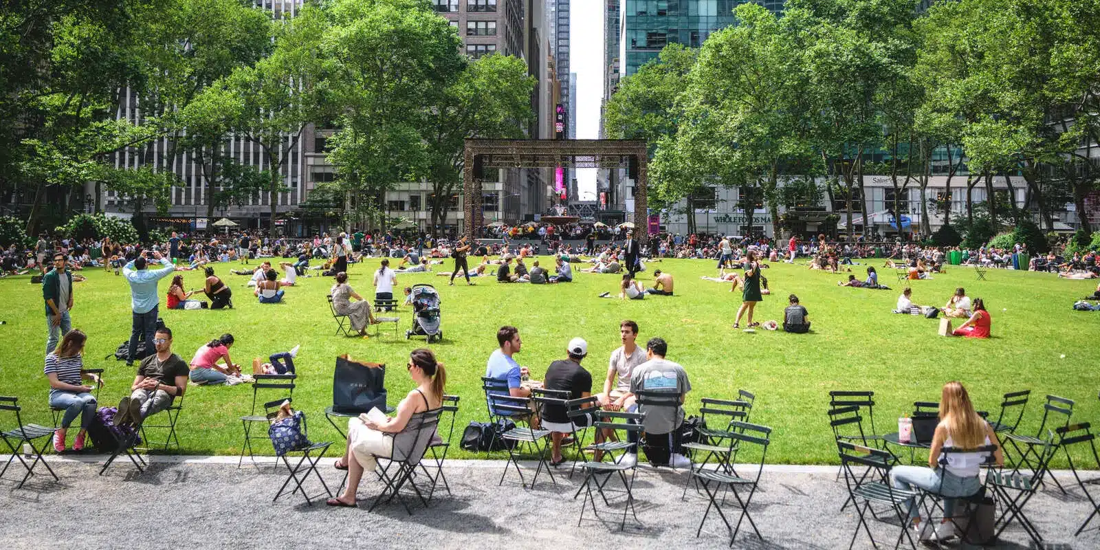 Crowd gathers on the lawn at Bryant Park in NYC, enjoying a sunny day.