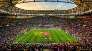 Aerial view of World Cup stadium with illuminated pitch and colorful crowd during golden hour