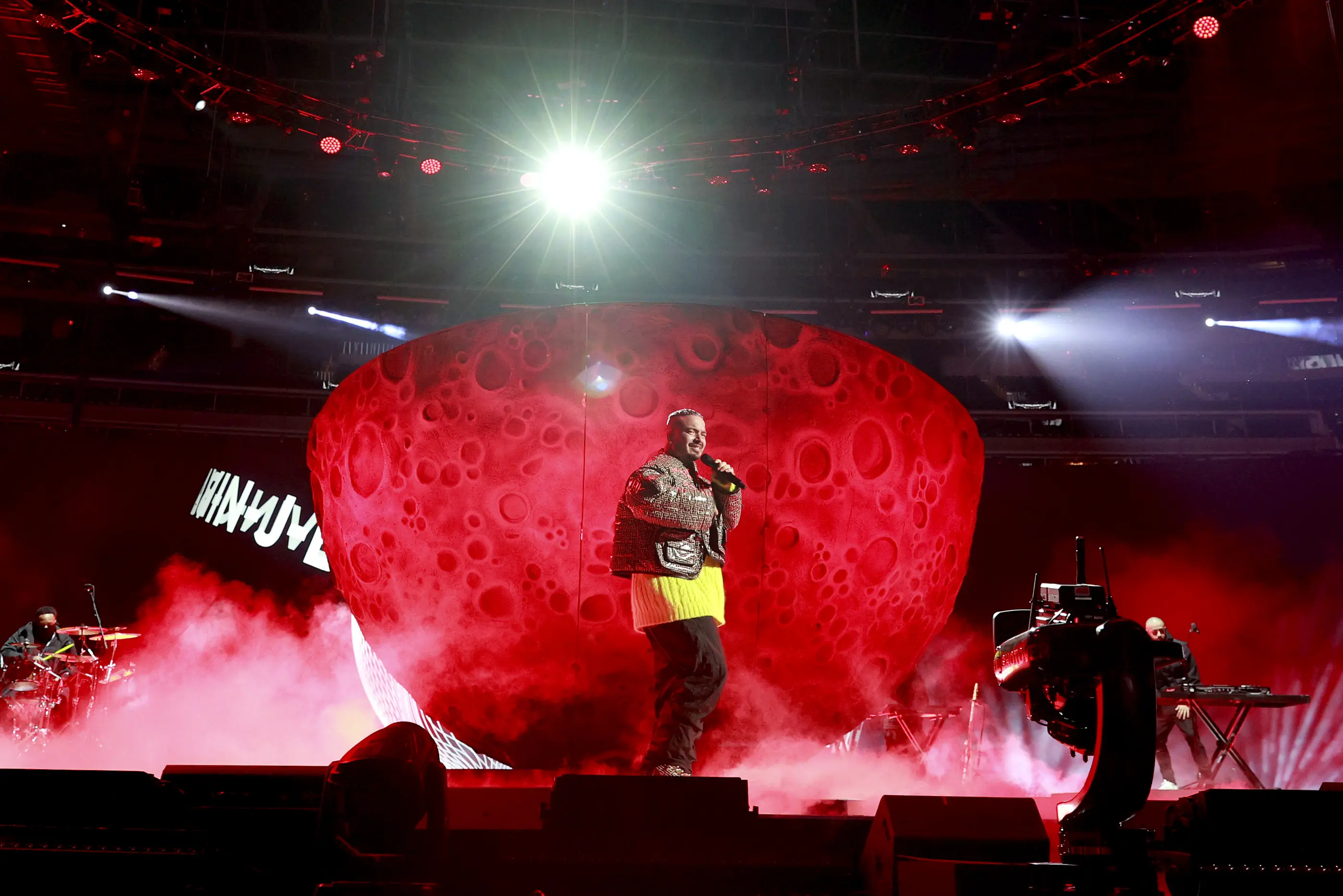 J Balvin performing on stage with dramatic lighting and a large red backdrop.