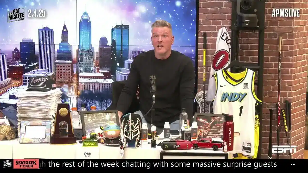 Pat McAfee in front of a studio desk with various sports memorabilia and a city backdrop.