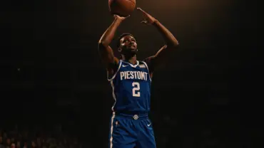 Basketball wing player shooting three-pointer in Detroit Pistons uniform with dramatic arena lighting