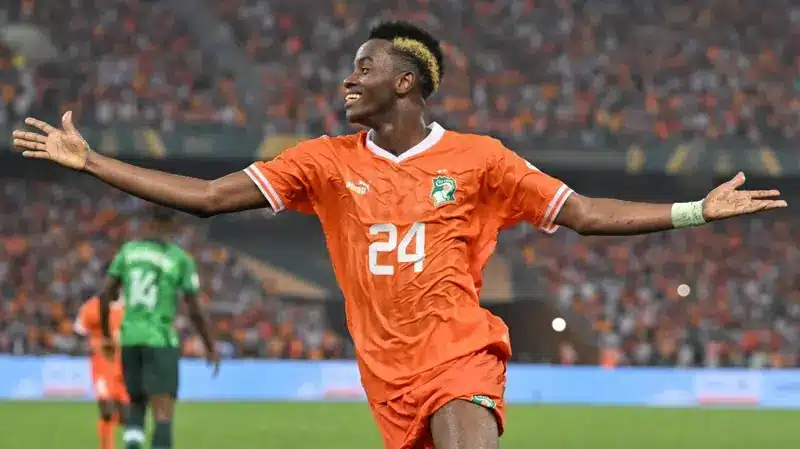 Simon Adingra celebrating a goal in an orange Ivory Coast football kit.