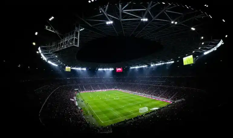 Illuminated soccer stadium at night with players on the field and a cheering crowd.