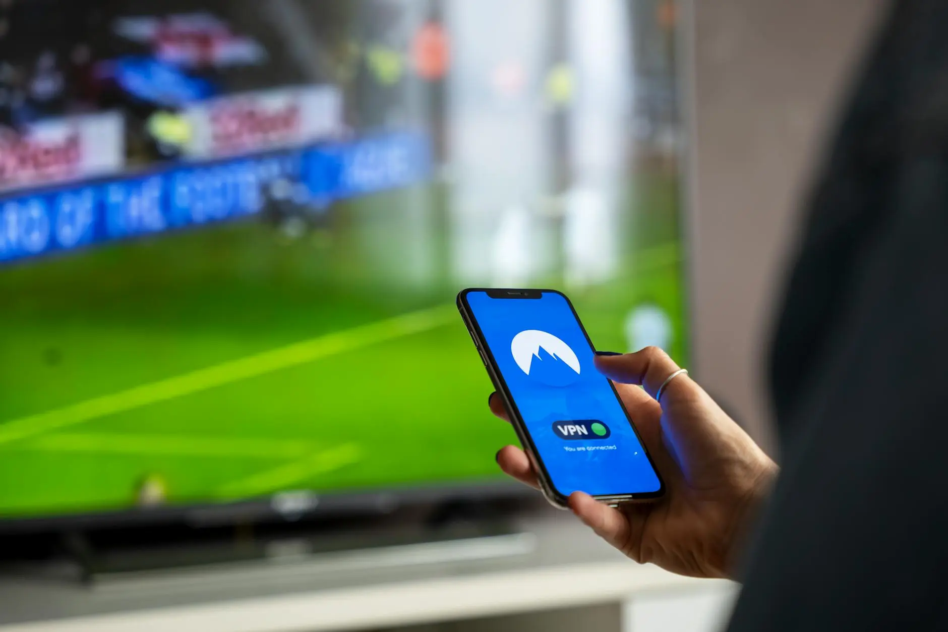Person holding a smartphone with a VPN app open, watching sports on TV.