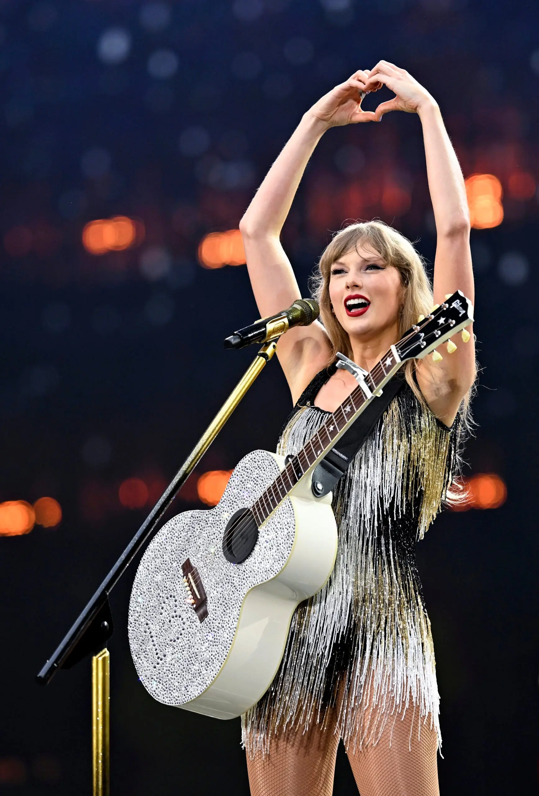Taylor Swift performing on stage with a white glittery guitar and making a heart gesture.