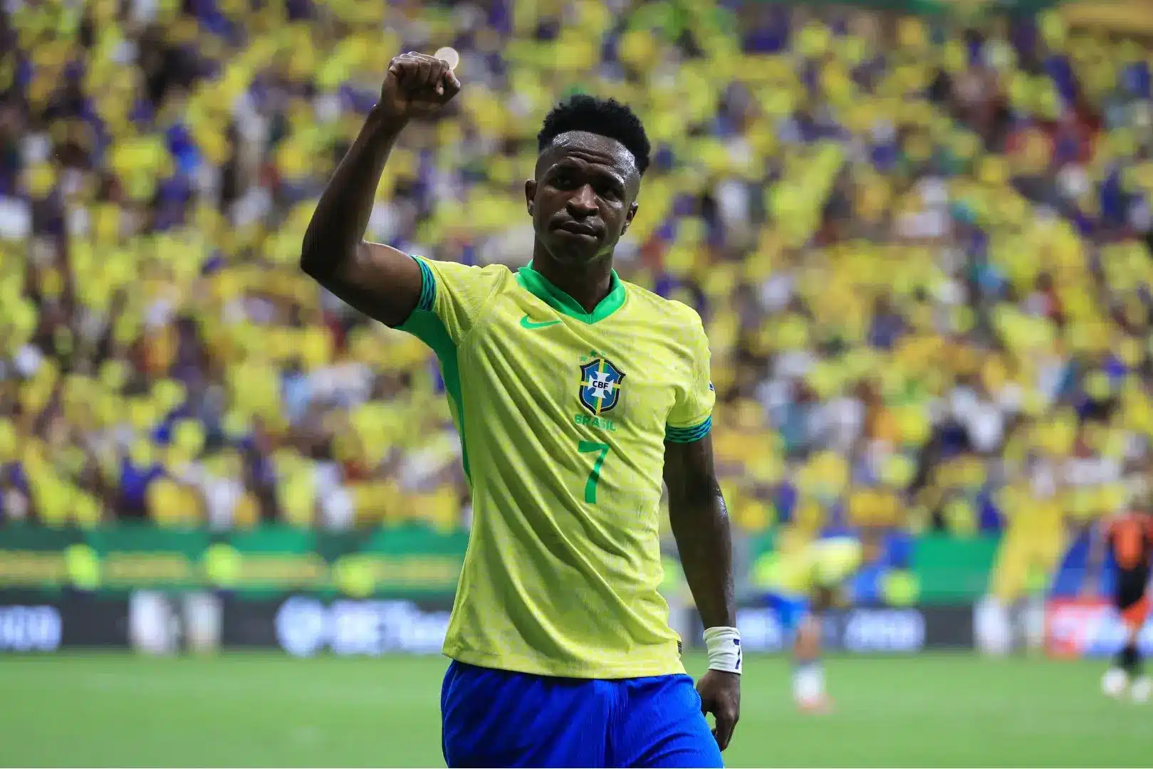 Vinicius Junior celebrates a goal in Brazil's national team uniform with a cheering crowd in the background.