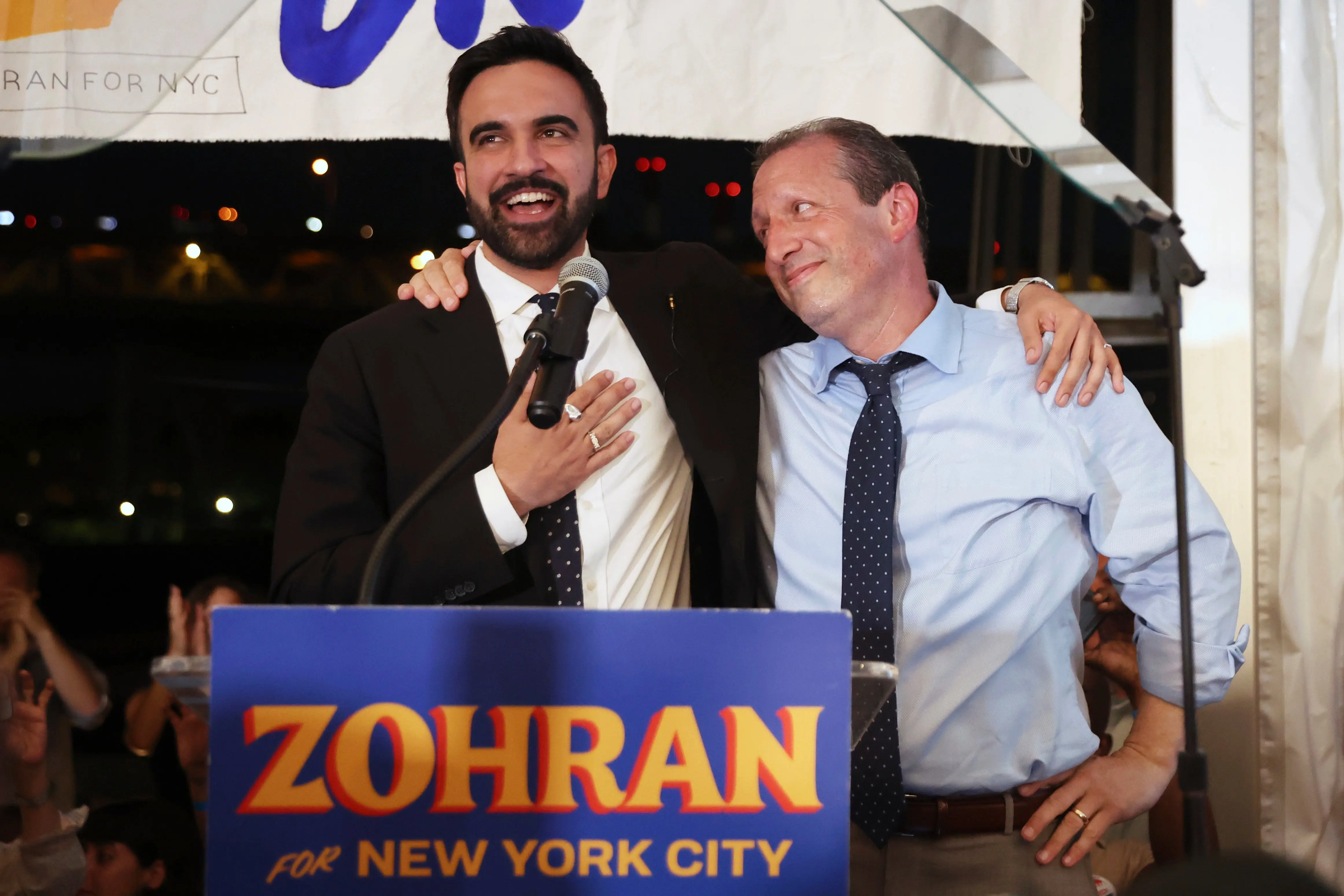 Zohran Mamdani speaking at an event with a supporter, both smiling joyfully.