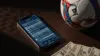 Smartphone showing sports betting parlay slip beside World Cup soccer ball and cash on dark table