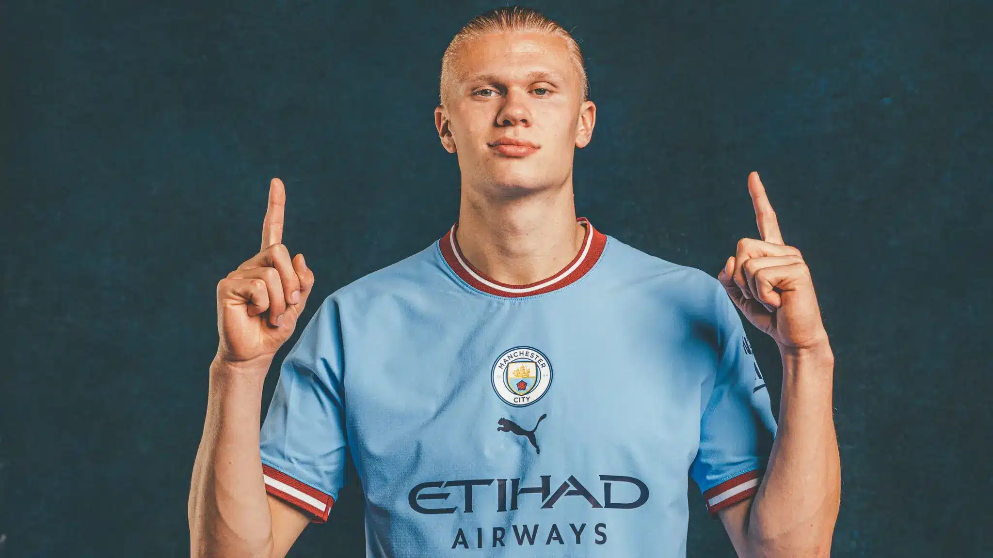 Erling Haaland in Manchester City jersey, raising both hands with index fingers pointing up.