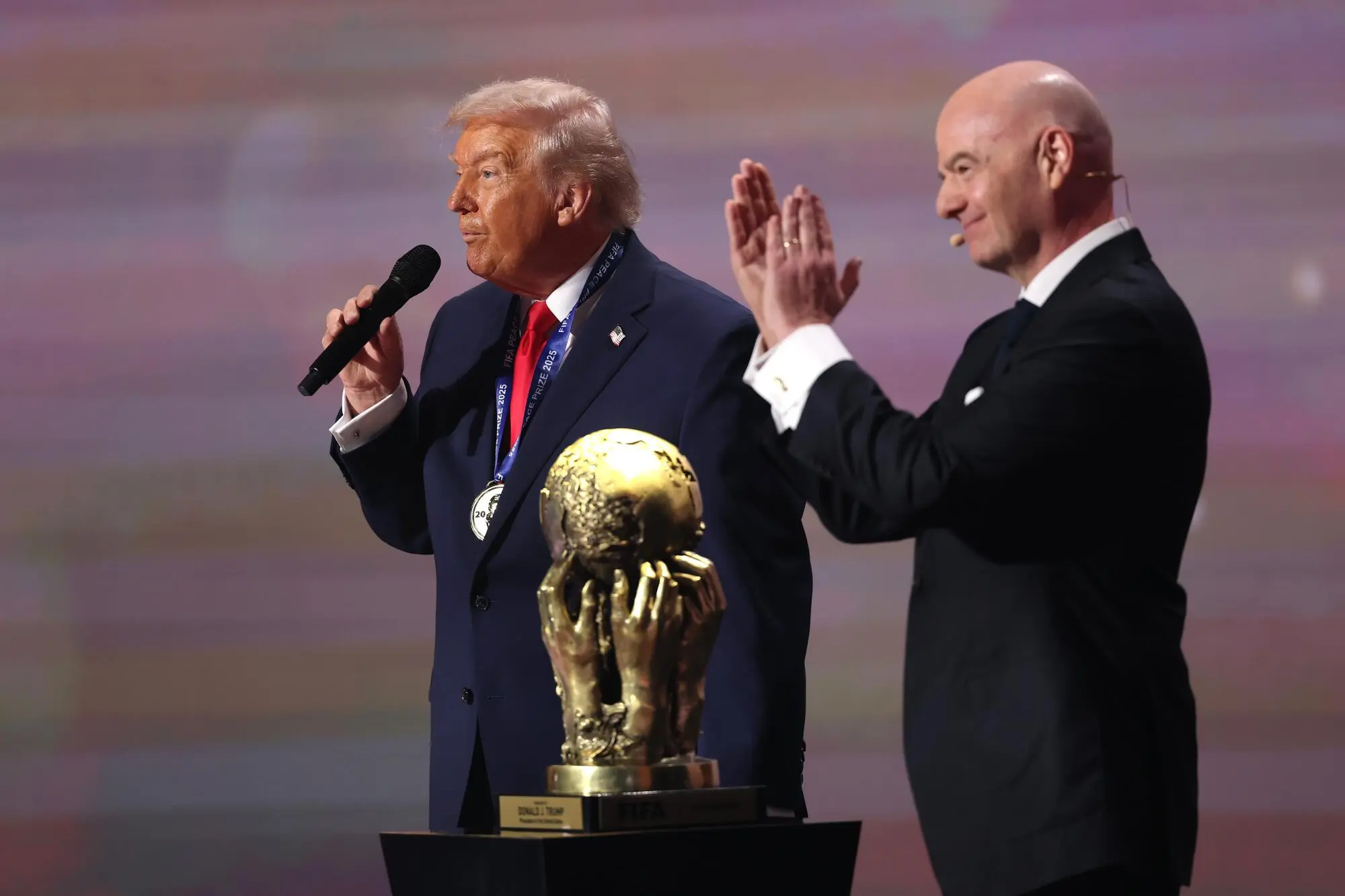 Donald Trump speaking into a microphone with Gianni Infantino applauding beside the FIFA trophy.