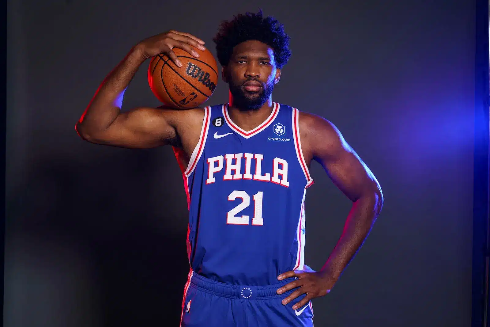 Joel Embiid wearing a blue 76ers jersey, holding a basketball.