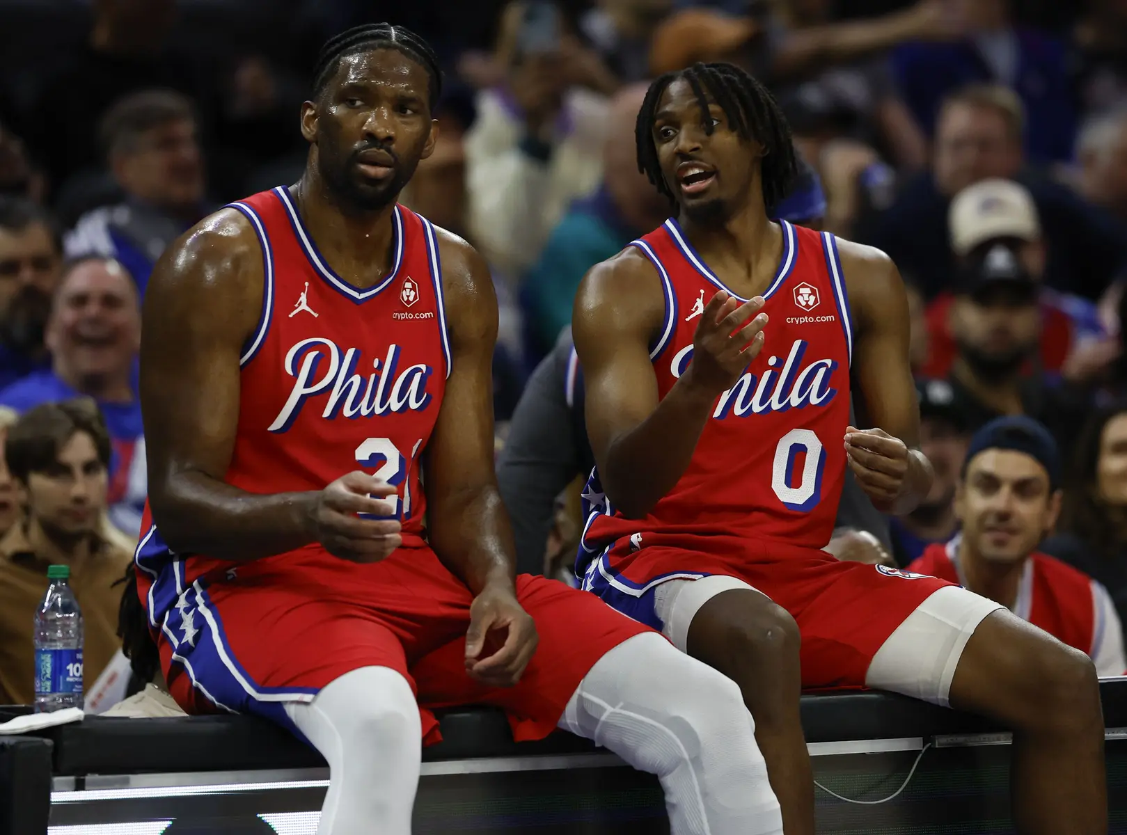 Joel Embiid and Tyrese Maxey in red 76ers jerseys seated on the bench during a game.