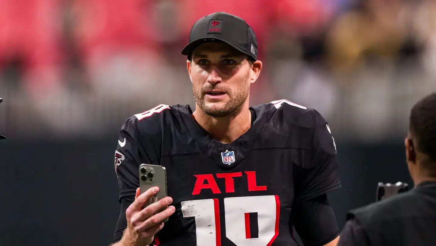 Quarterback wearing Falcons jersey and cap, holding a phone on the field.