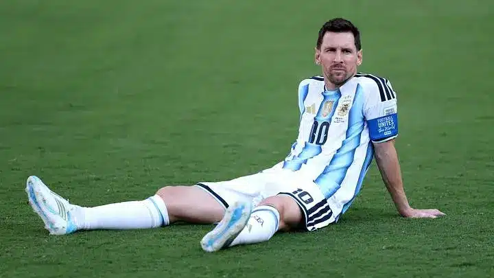 Lionel Messi wearing Argentina's jersey, sitting on the field.
