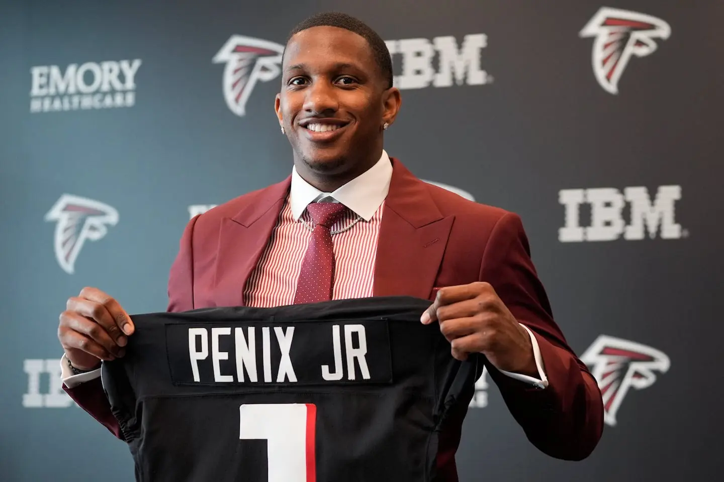 Michael Penix Jr. wearing a red suit and holding up an Atlanta Falcons jersey with his name and number 1.