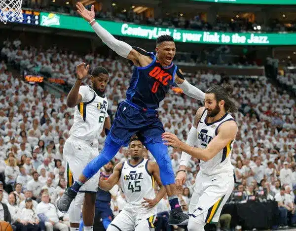 Russell Westbrook jumps over defenders during an NBA game with fans in the background.