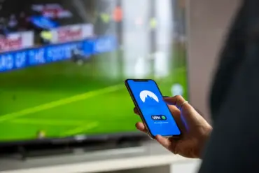Person holding smartphone displaying a VPN app with a sports game on TV in the background.