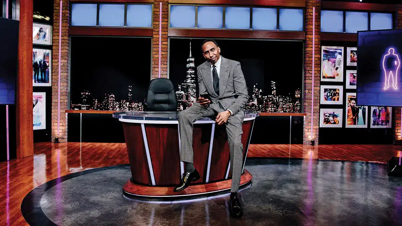 A man in a suit sitting on a desk in a television studio with a city skyline backdrop.