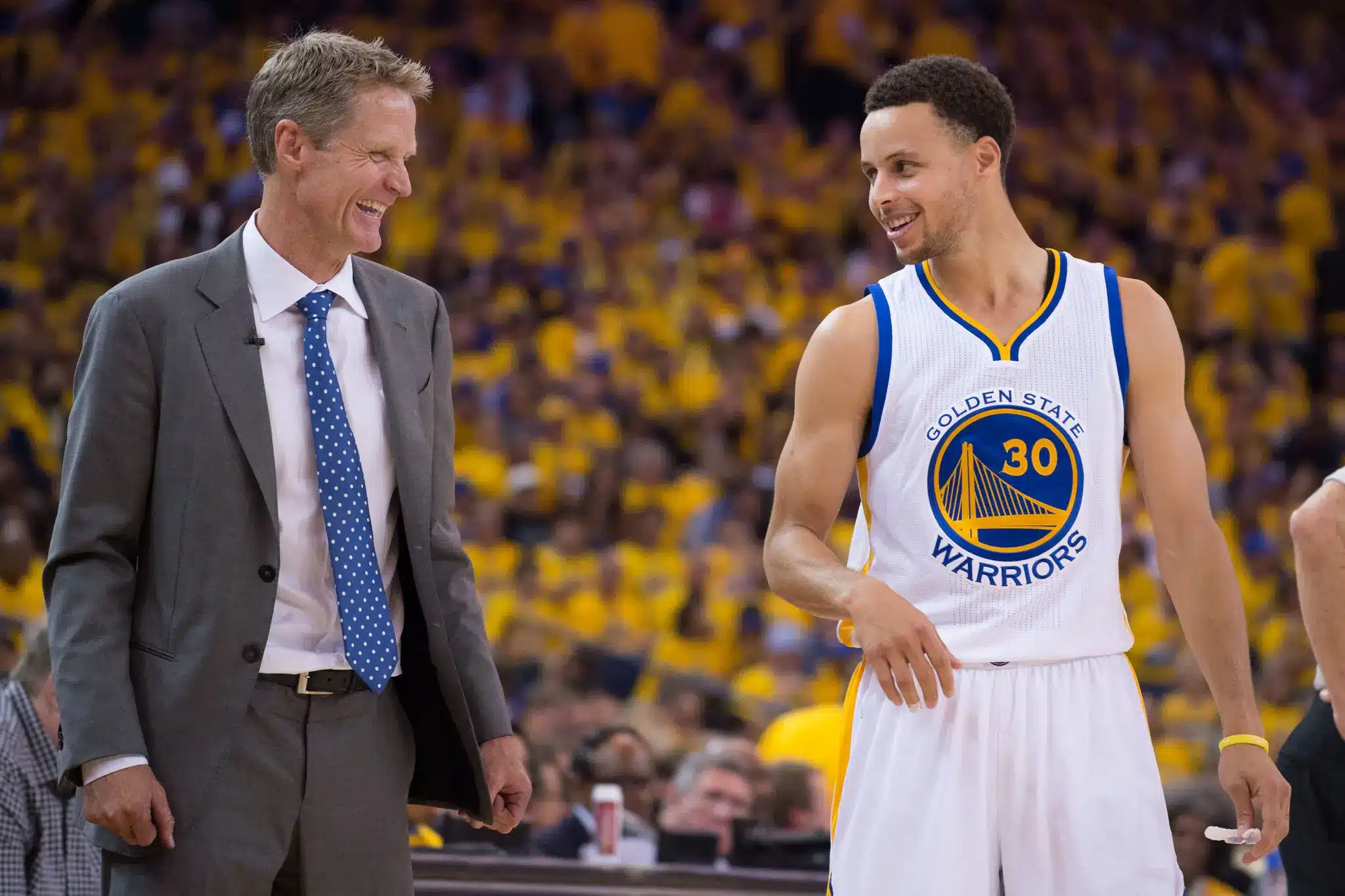 Steve Kerr and Stephen Curry smiling during a Golden State Warriors game.