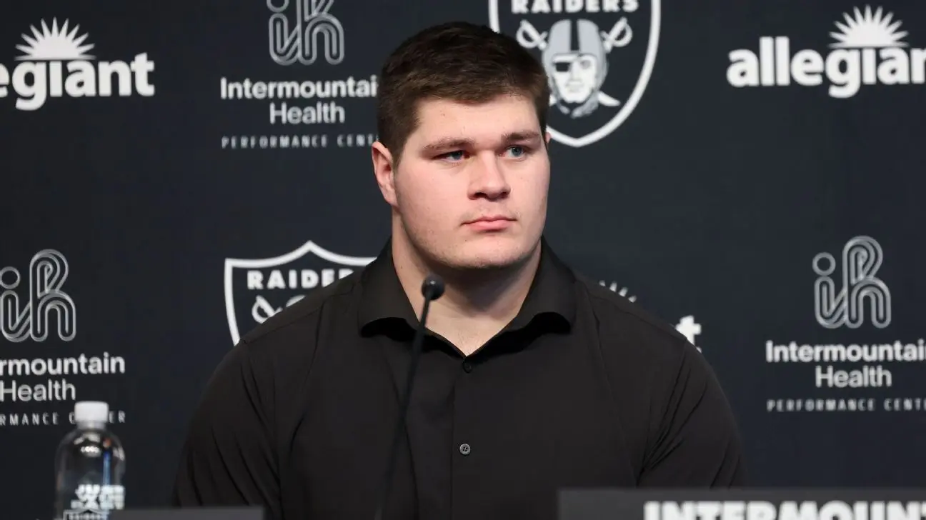 Tyler Linderbaum seated at a press conference with Raiders branding in the background.