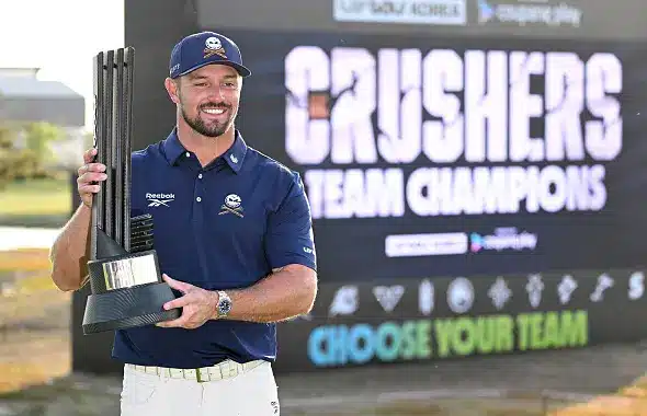 Bryson DeChambeau holding a trophy in front of a Crushers Team Champions backdrop