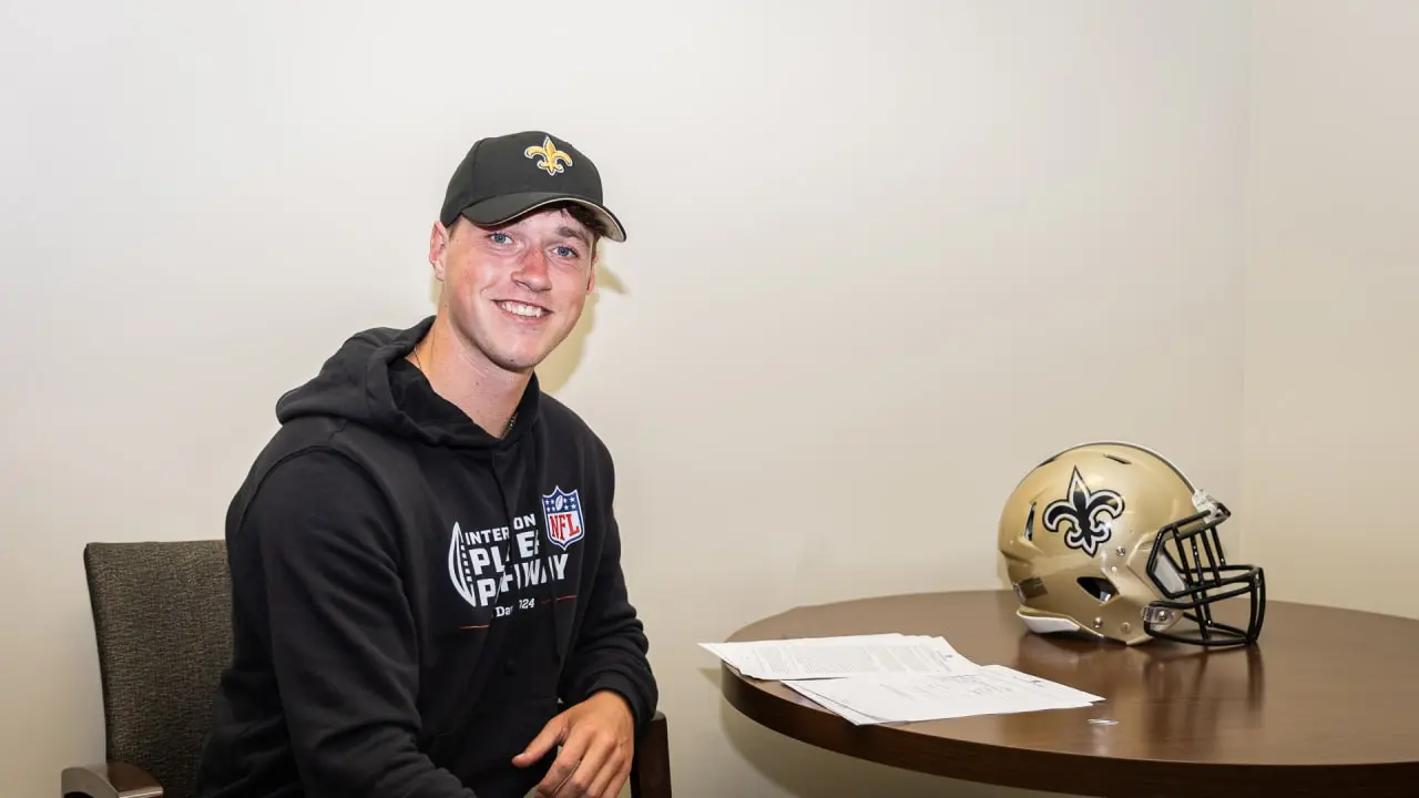 Charlie Smyth wearing a New Orleans Saints hat and hoodie sitting next to a Saints helmet on a table