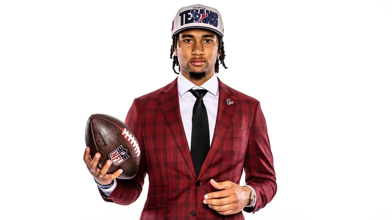 CJ Stroud wearing a red plaid suit and a Houston Texans hat, holding an NFL football