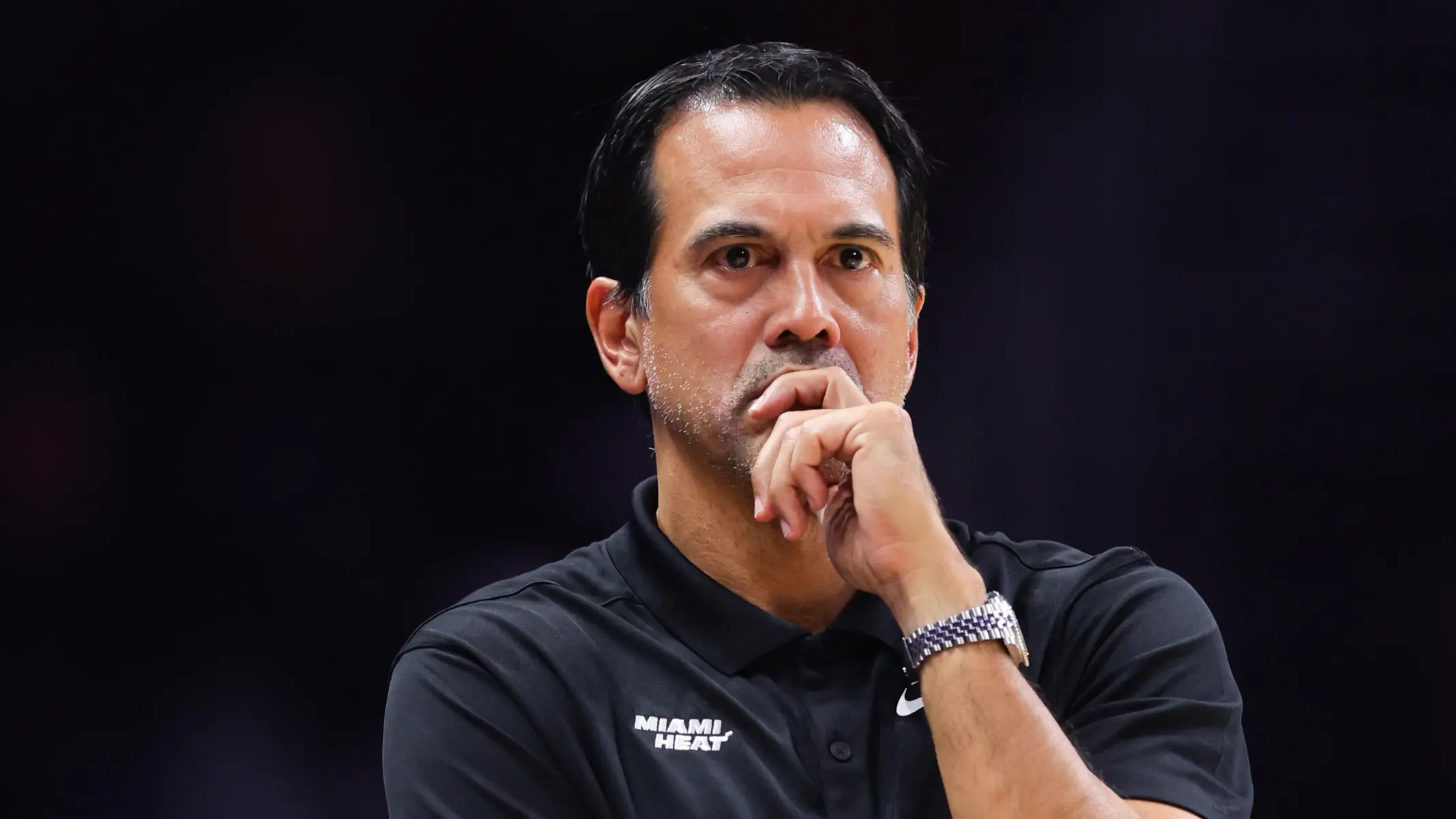 Miami Heat coach Erik Spoelstra wearing a black team polo shirt, looking thoughtful with hand on chin.