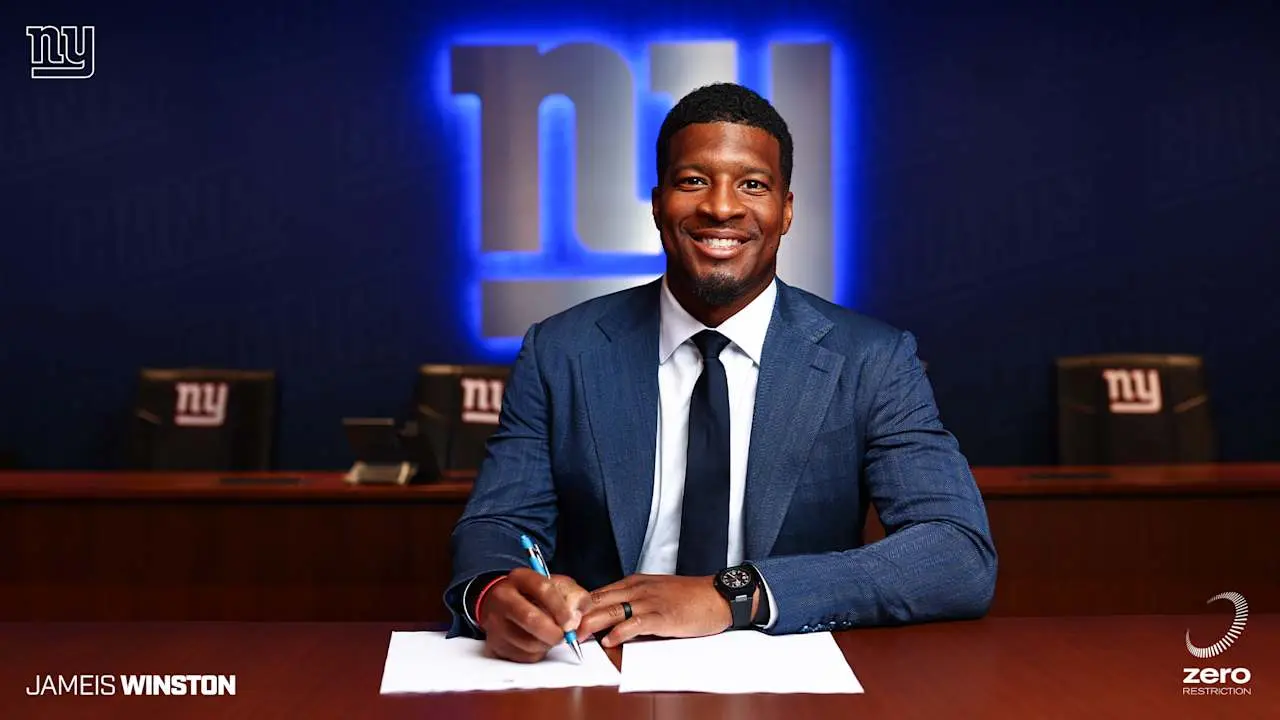 Jameis Winston wearing a blue suit and tie, signing a contract in front of New York Giants logos.