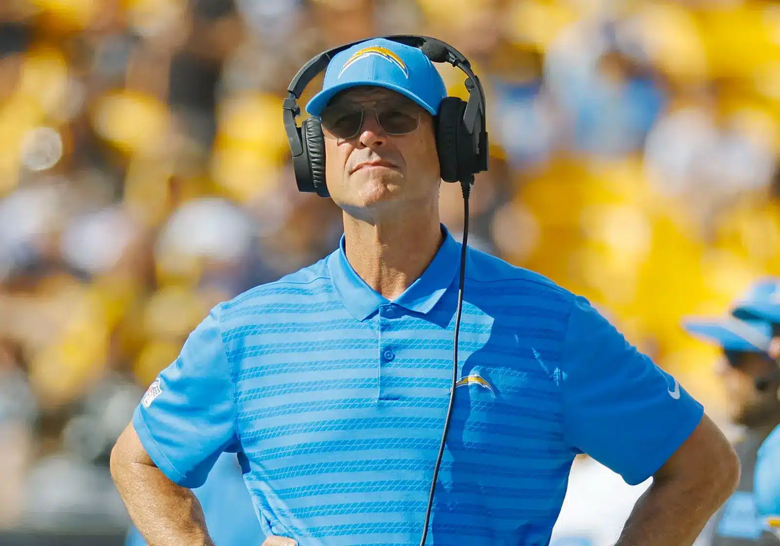 Jim Harbaugh wearing a blue Los Angeles Chargers polo, hat, and headset on the sidelines.