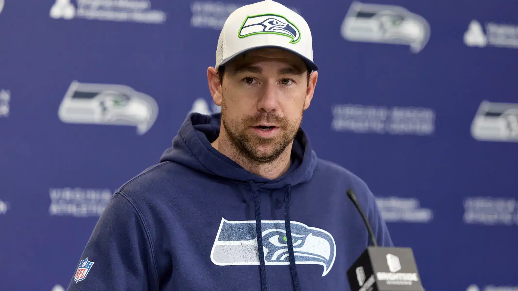Klint Kubiak wearing a Seattle Seahawks hat and hoodie speaking at a press conference