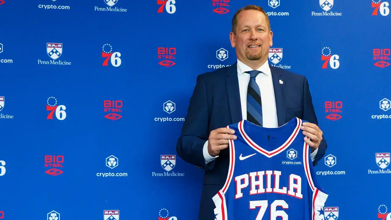 Nick Nurse wearing a navy suit and holding up a Philadelphia 76ers jersey in front of a team backdrop