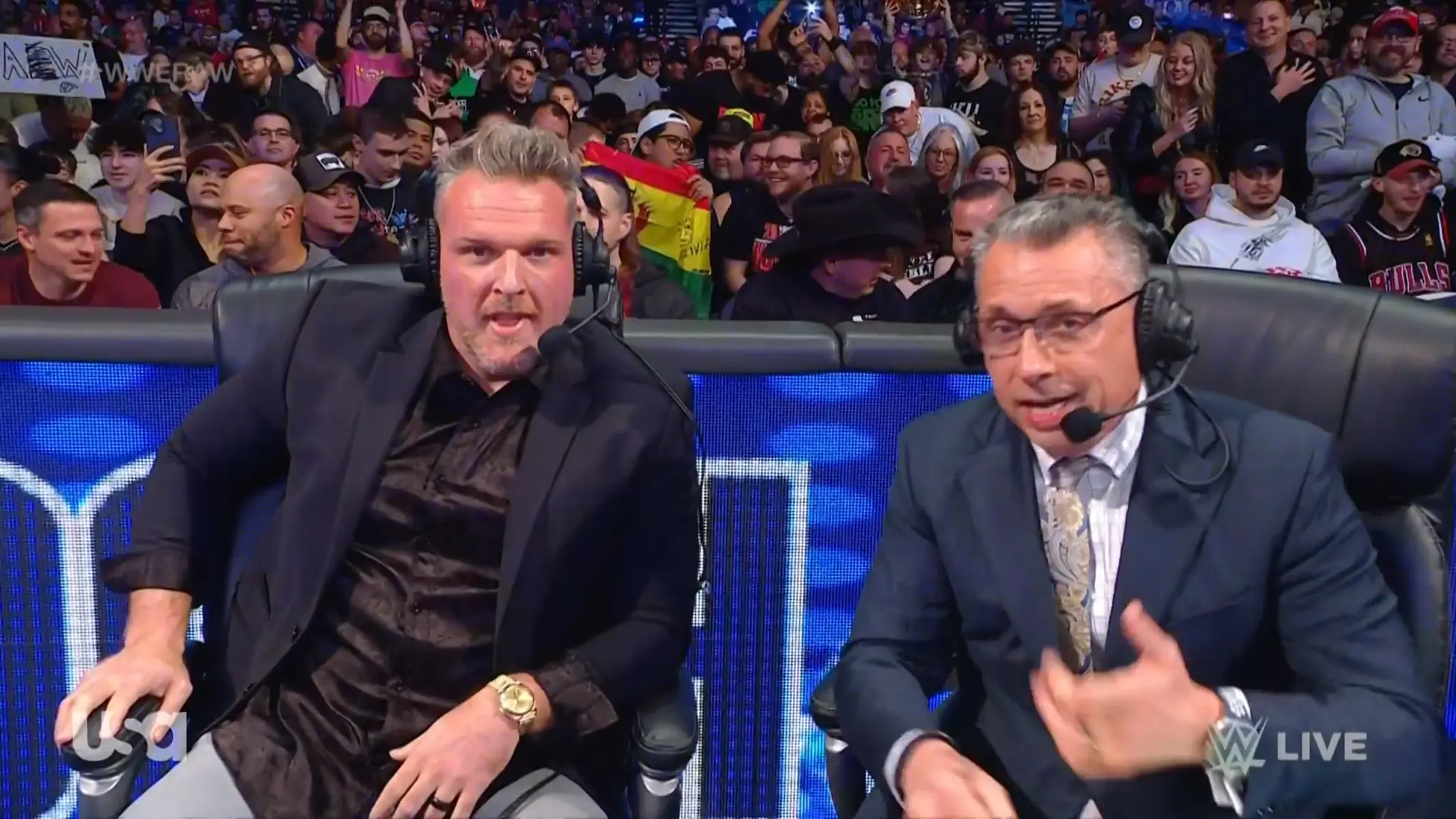 Pat McAfee and Michael Cole wearing headsets at a WWE commentary table during a live event.